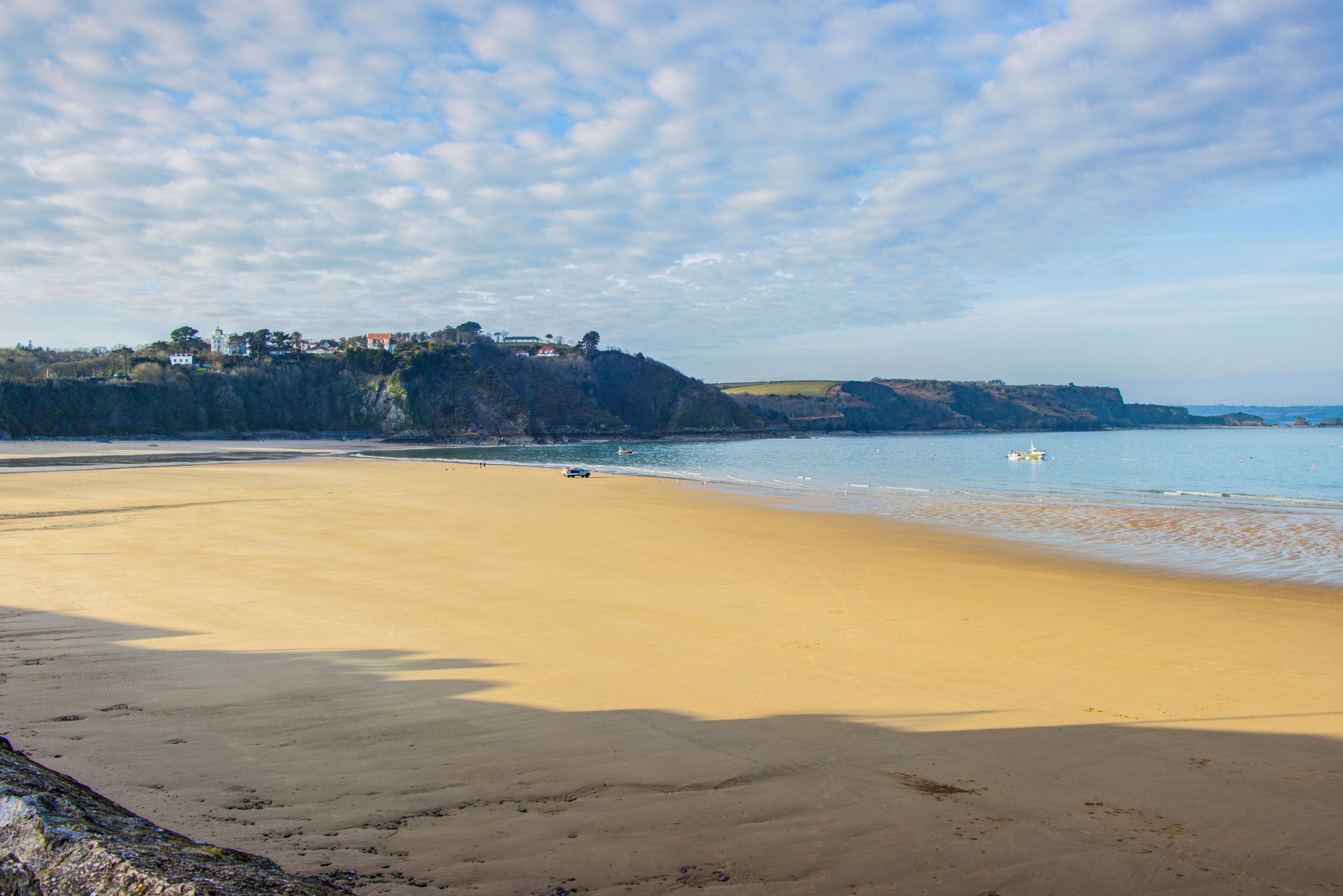 A wide sandy beach with a calm sea, a distant hill with buildings, and a clear sky.