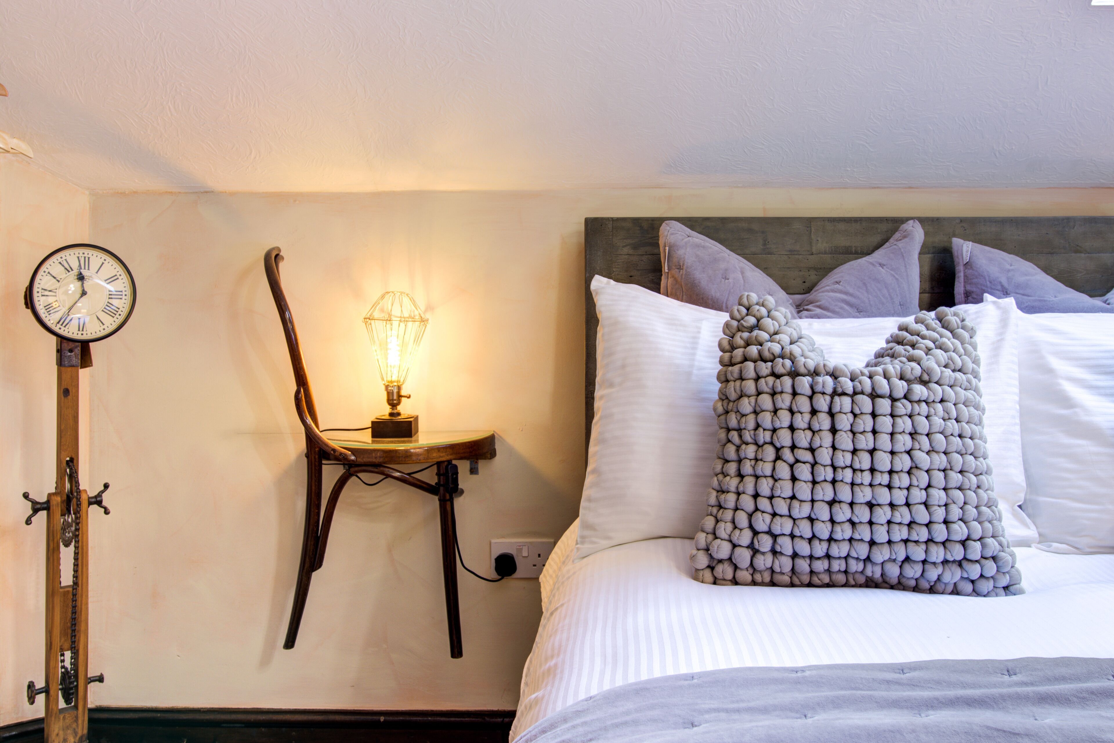A neatly made bed with a unique textured pillow, a wall-mounted clock, and a small table with a lamp.