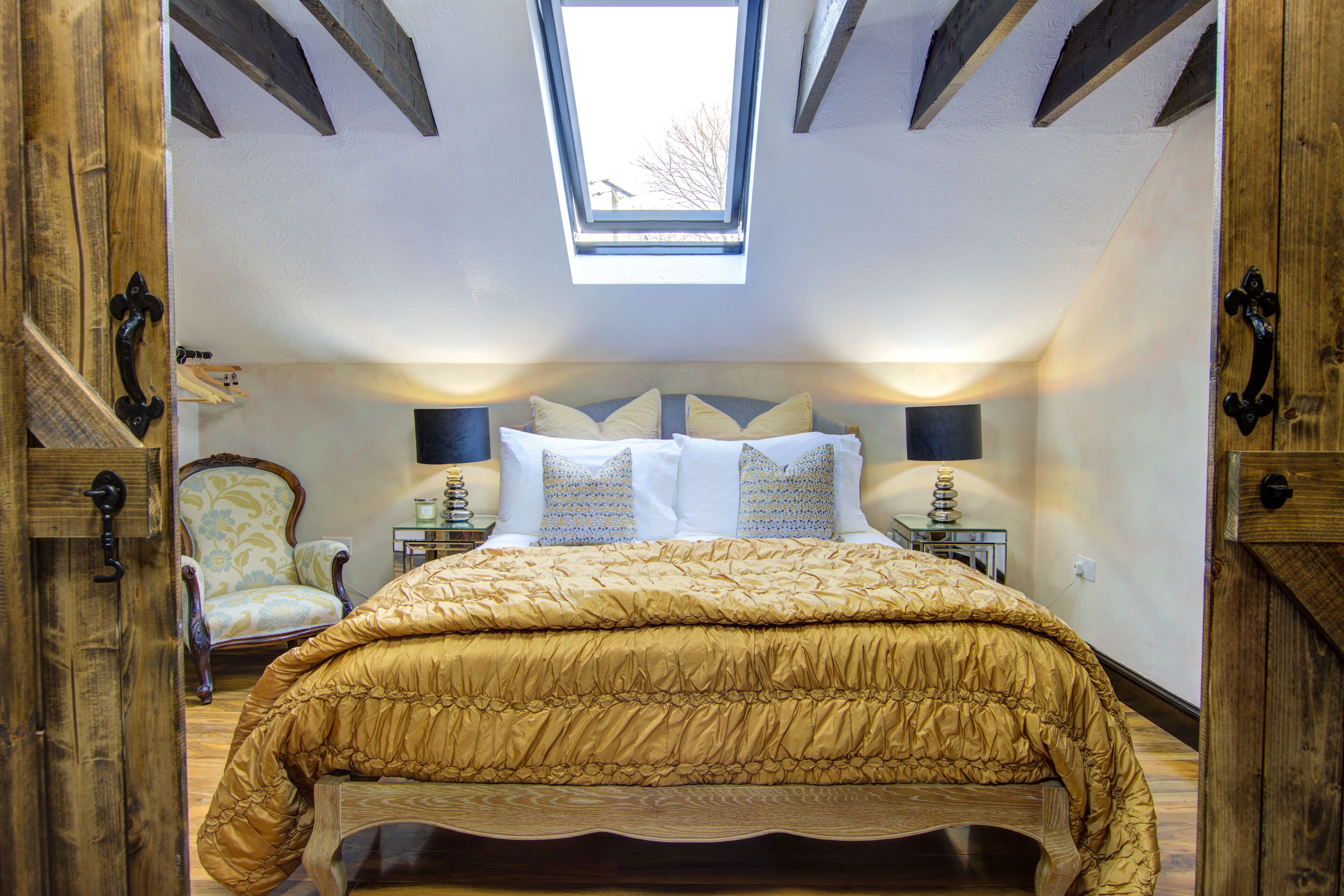 A bedroom with a vaulted ceiling, a skylight, a bed with a gold quilt, two bedside tables with lamps, and a chair.