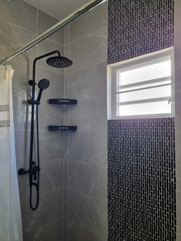A shower area with a rain showerhead, handheld showerhead, and a handheld massager. There is a window with horizontal blinds and a tiled wall with a brick pattern.