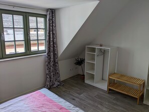 Une chambre à coucher avec un lit, une fenêtre avec des rideaux, une armoire blanche, un banc en bois et une plante.