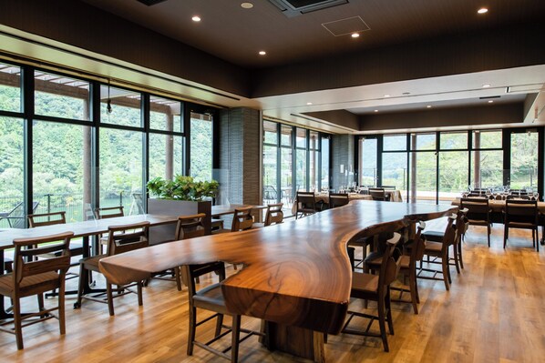 A spacious conference room with a long wooden table, multiple chairs, large windows, and a view of greenery outside.