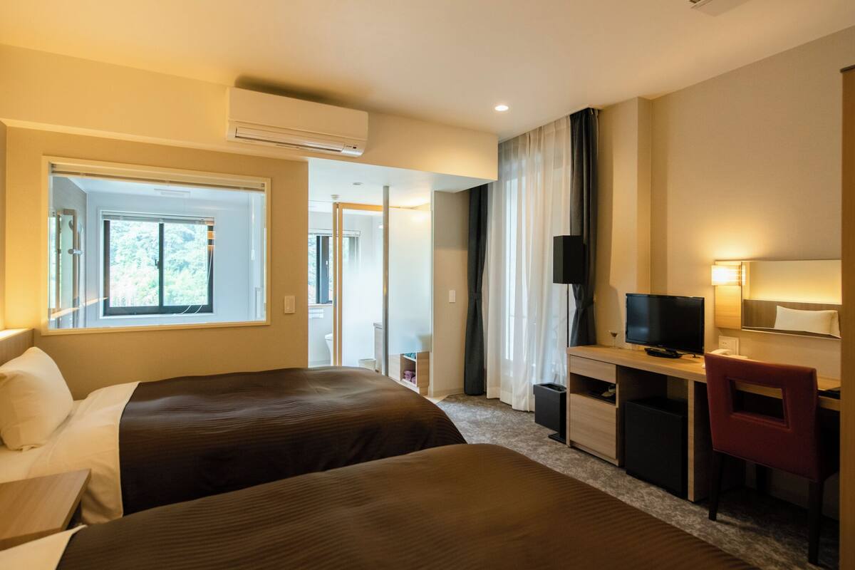 A hotel room with two beds, a desk, a TV, and an air conditioning unit.