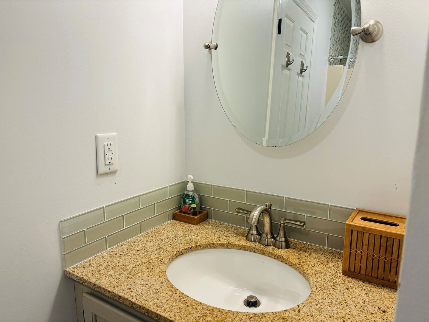 Une salle de bain avec un lavabo doté d’un plan de travail en granit, un miroir, un distributeur de savon et une boîte à mouchoirs en bois.