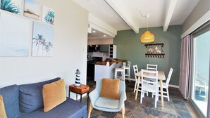 A living room with a sofa, armchairs, a dining table with chairs, a kitchen in the background, and artwork on the walls. - Aquarius Condo Unit 207 2 Bedroom Condo by RedAwning (South Padre Island)