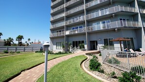 A multi-story residential building with balconies, a paved walkway, and a small outdoor seating area. - Aquarius Condo Unit 207 2 Bedroom Condo by RedAwning (South Padre Island)