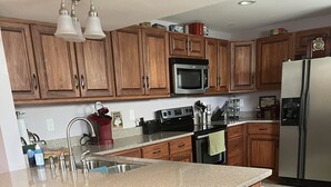 Eine Küche mit Holzausbau, einem Edelstahlkühlschrank, einer Mikrowelle und einer Arbeitsplatte aus Granit. Es gibt drei Barhocker und eine Spüle mit Wasserhahn.