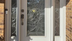 Eine weiße Doppeltür mit einem durchscheinenden Glasfeld mit Blattform-Motiv, eingelassen in eine Ziegelwand.