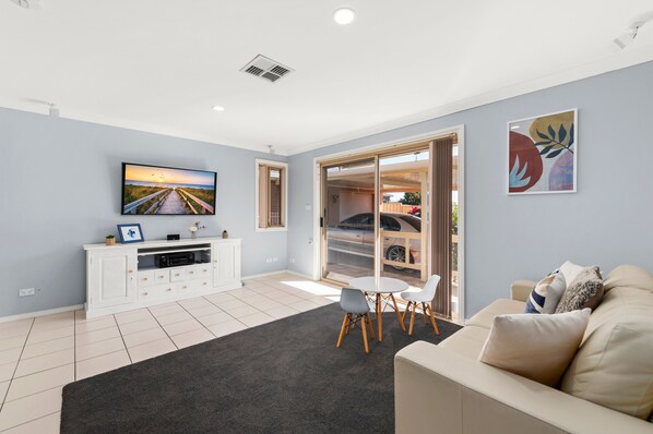 A modern living room with a flat-screen TV, a white entertainment unit, a grey sofa, and a dining area with a table and chairs.