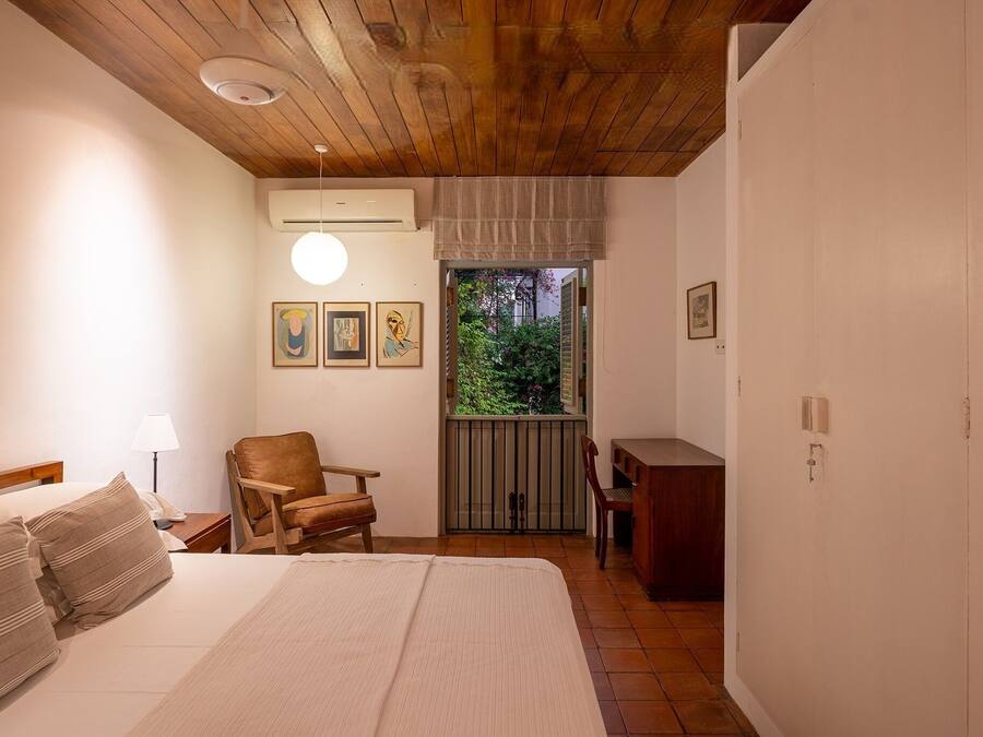 A bedroom with a bed, a chair, a desk, and a window with a view of greenery.