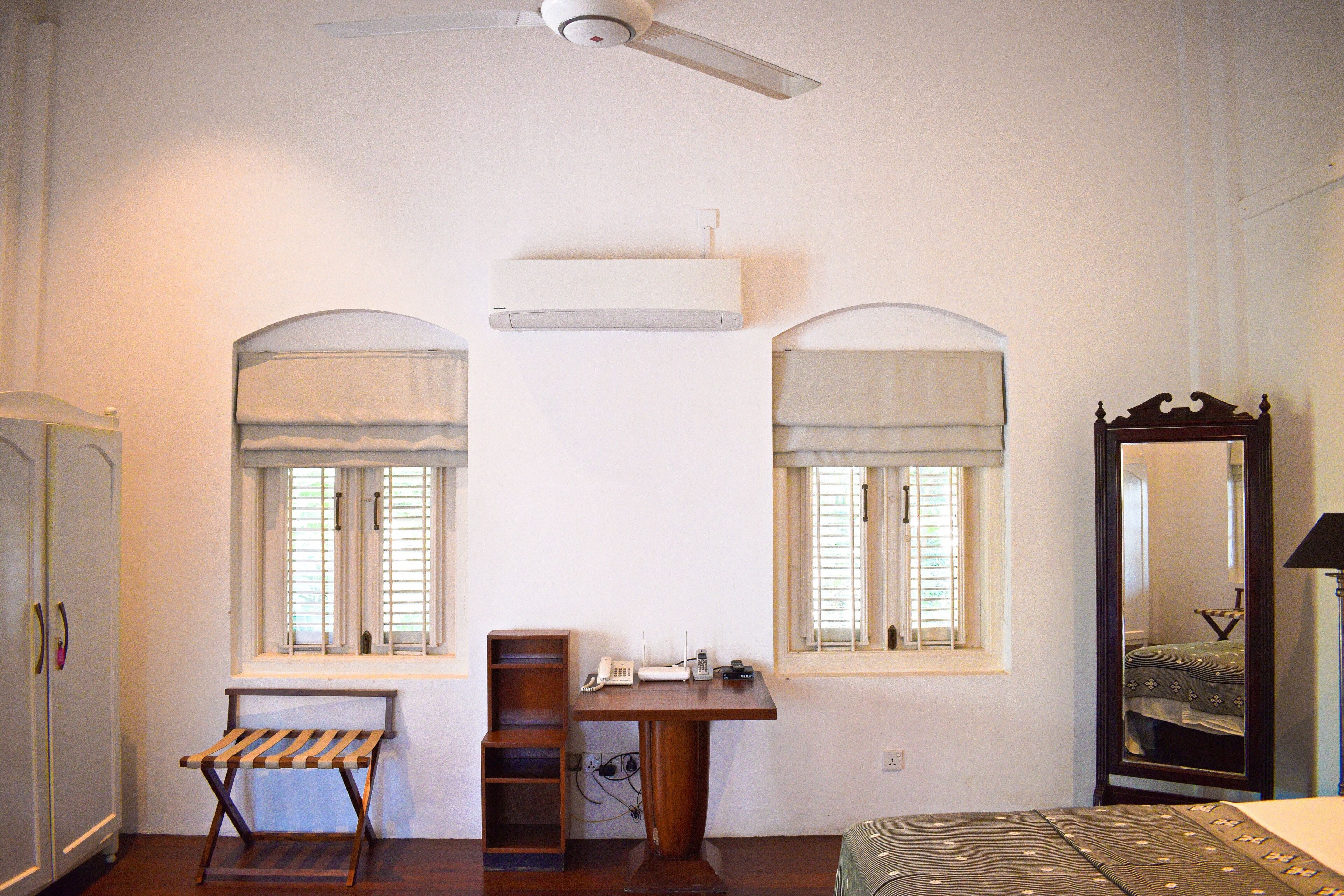 A bedroom with a bed, a desk, two windows with curtains, a wardrobe, and a ceiling fan.