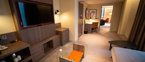 Een hotelkamer met een flatscreen televisie, een glazen salontafel, een bank, een bureau en een stoel.