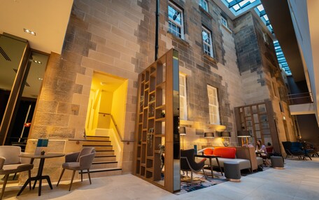 A spacious indoor area with a high ceiling, stone walls, and a staircase leading to an upper level. There are seating areas with tables and chairs, and a small table with a plant.