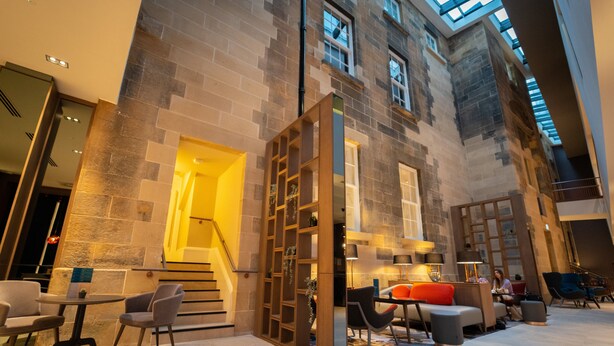 A spacious indoor area with a high ceiling, stone walls, and a staircase leading to an upper level. There are seating areas with tables and chairs, and a small table with a plant.