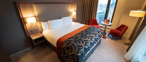 A hotel room with a large bed, a bedside table with a lamp, a small table with a bottle, a red chair, and a view of the city through a window.