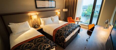 A hotel room with two beds, a desk, a chair, and a window with a view of buildings and greenery.
