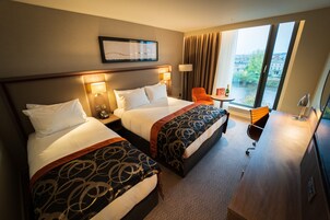 Habitación de hotel con dos camas, un escritorio, una silla y un ventanal con vistas a edificios y áreas verdes.