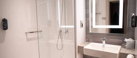 Un baño moderno con ducha de vidrio, un espejo grande con iluminación integrada, un lavamanos blanco y un perchero para toallas.