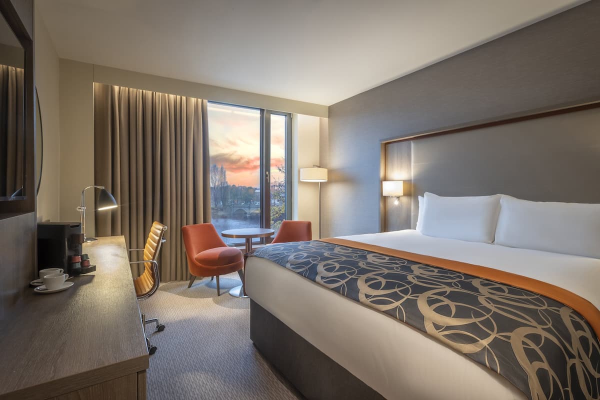 Une chambre d’hôtel avec un grand lit, un bureau avec une lampe, une chaise et une vue sur un coucher de soleil au-dessus d’un pont.