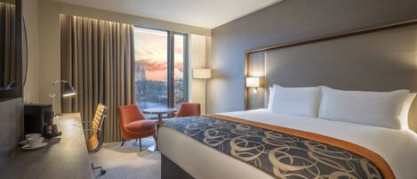 A hotel room with a large bed, a desk with a lamp, a chair, and a view of a sunset over a bridge.