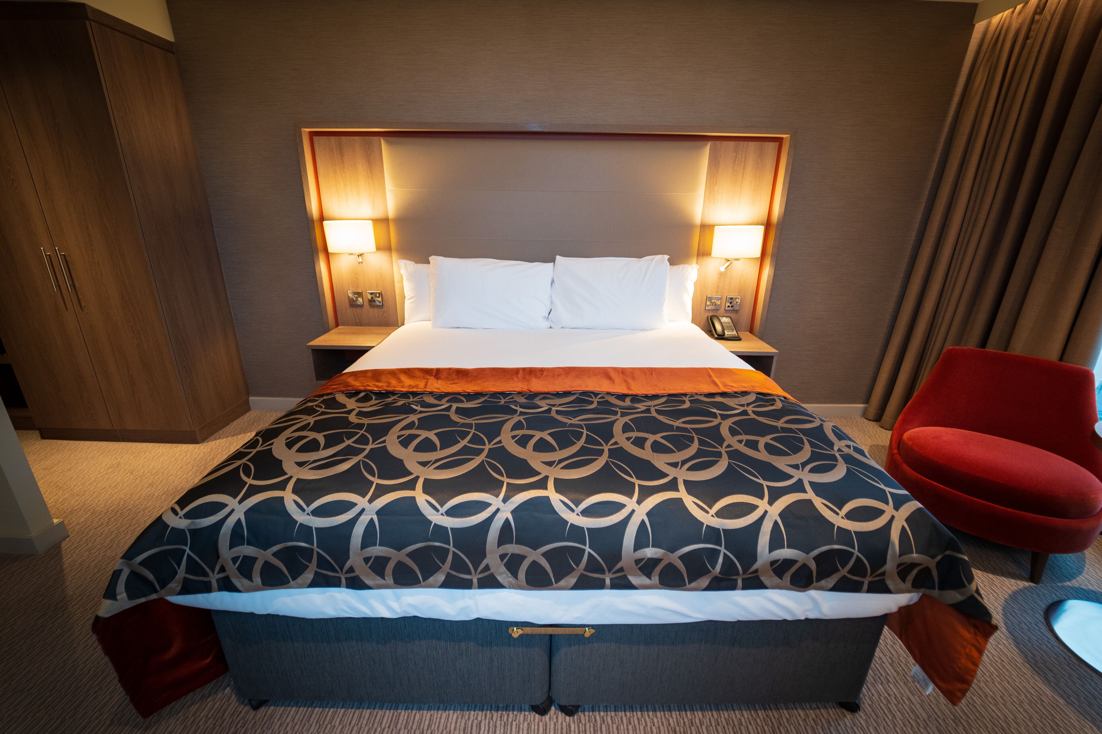 A hotel room with a large bed, two bedside lamps, a red chair, and a wooden wardrobe.