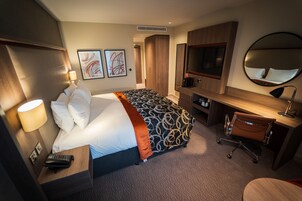 Habitación de hotel con cama, escritorio, silla, televisión, espejo y obras de arte en la pared.