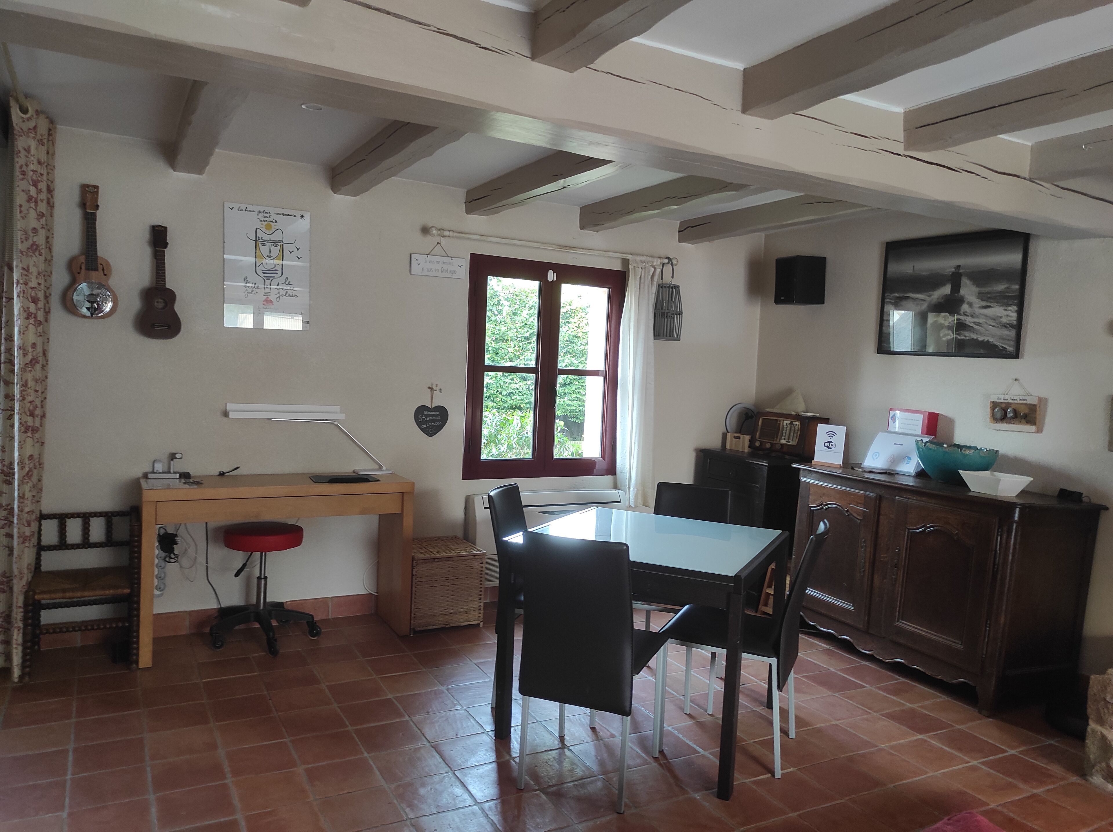 Une pièce comprenant un bureau en bois, une table à manger avec des chaises, un radiateur, une fenêtre avec des rideaux et une radio fixée au mur.