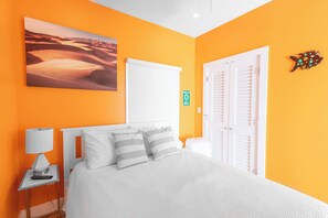 A bedroom with an orange wall, a bed with white bedding, a nightstand with a lamp, a painting of a desert landscape, and a door with a sign indicating the room's location. - Keys Breeze - Walk to Duval, Private Pool (Key West)