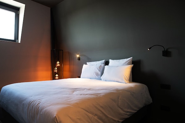 Una cama bien tendida con sábanas blancas en una habitación moderna con una pared oscura y una ventana a la izquierda.