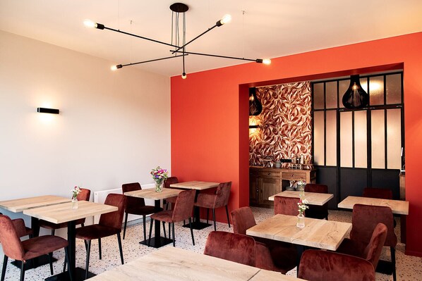 A modern dining area with wooden tables and red upholstered chairs, a decorative wall with a leaf pattern, and a unique ceiling light fixture. - Hôtel de la Poste (Ellezelles)
