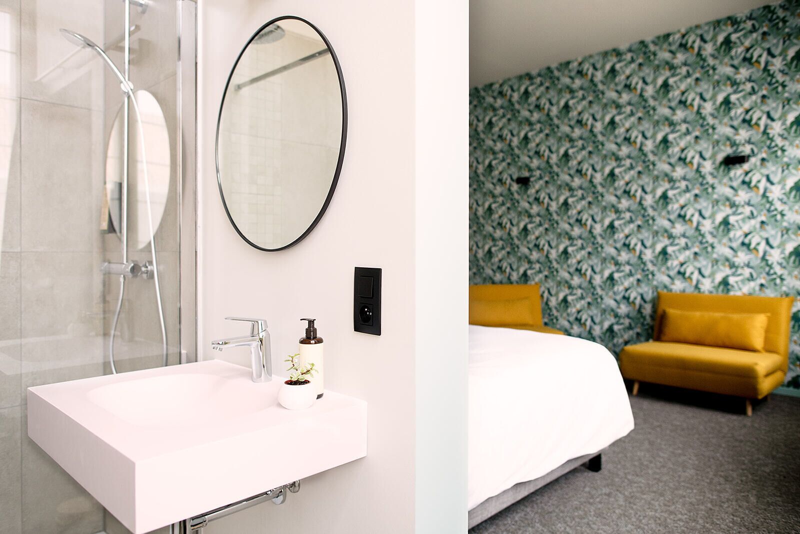 Un baño con un lavamanos blanco, un espejo y una ducha. La habitación tiene papel tapiz estampado y un sofá amarillo.