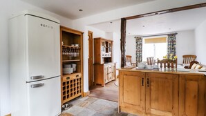 A kitchen with a white refrigerator, wooden cabinets, a dining table with chairs, and a window with curtains. - Anglers Barn (Skipton)