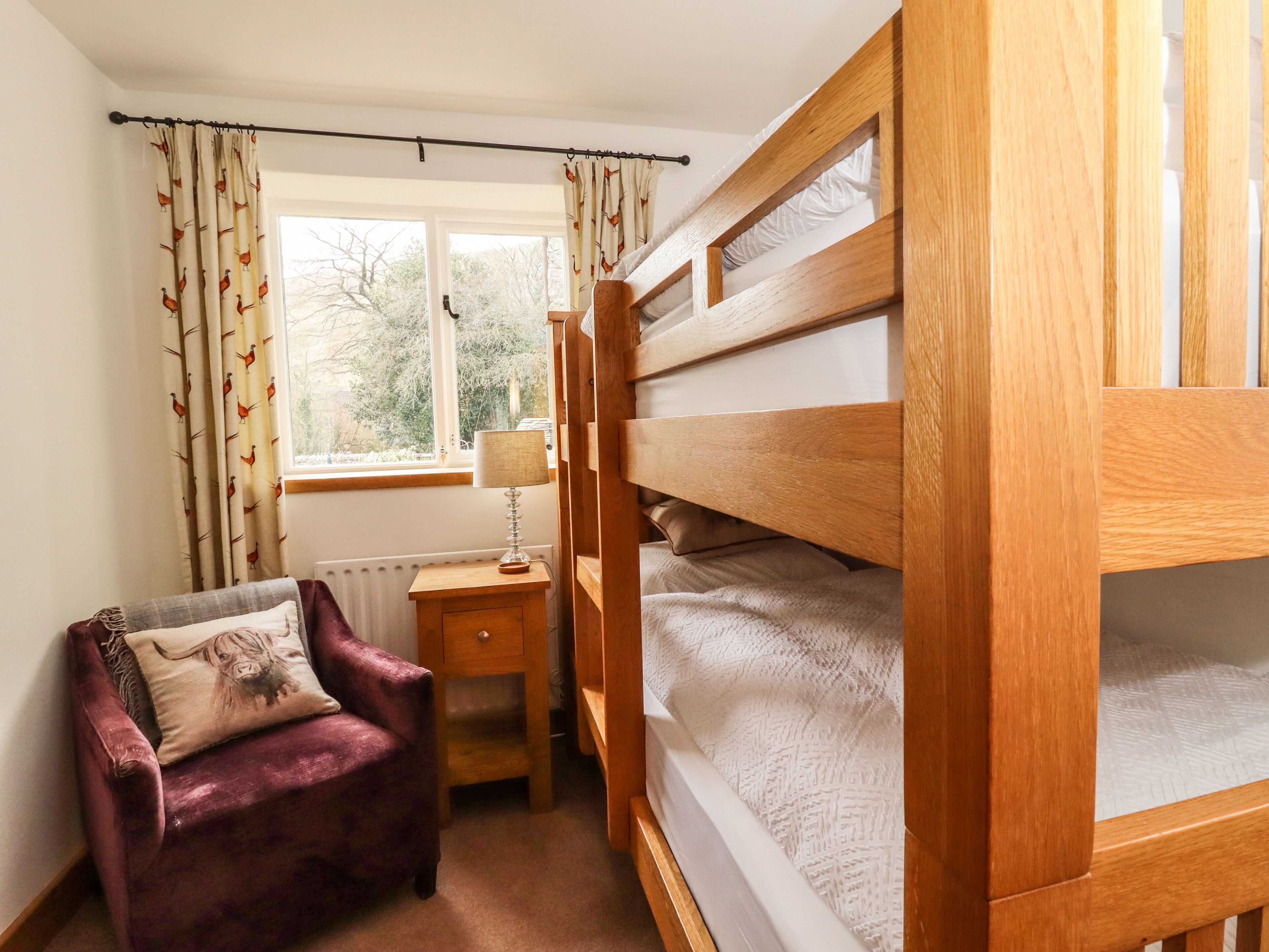 A bedroom with a bunk bed, a chair with a cushion, a bedside table with a lamp, and a window with curtains.