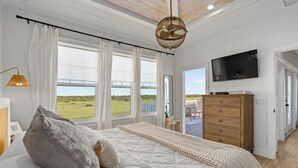 A bedroom with a large bed, a TV, a dresser, and a view of a green landscape through the windows. - Marsh front pool home with ocean, river and historical downtown views (St. Augustine)