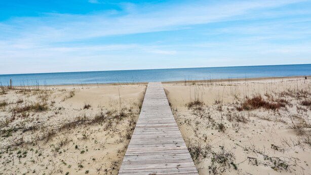 Une promenade en bois qui mène à une plage bordée de dunes de sable et d’une végétation clairsemée.