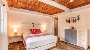 A bedroom with a wooden ceiling, a bed with white bedding, a bedside table with a lamp, and a white wardrobe.