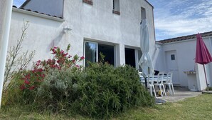 Un edificio blanco con un gran ventanal abierto, una ventana pequeña y un patio amueblado con sillas y una sombrilla.