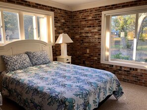Une chambre avec un mur en briques, un lit recouvert d’une literie à motifs floraux, une table de chevet avec une lampe et des fenêtres donnant sur des arbres.