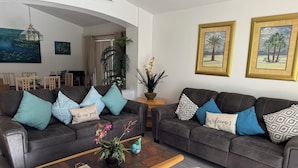 A modern living room with two dark brown sofas, each adorned with patterned cushions. A wooden coffee table with a vase of flowers is in the center. The room features a large window with curtains, a chandelier, and framed artwork on the walls. - Entire Villa with Pool and great Privacy near Disney (Kissimmee)