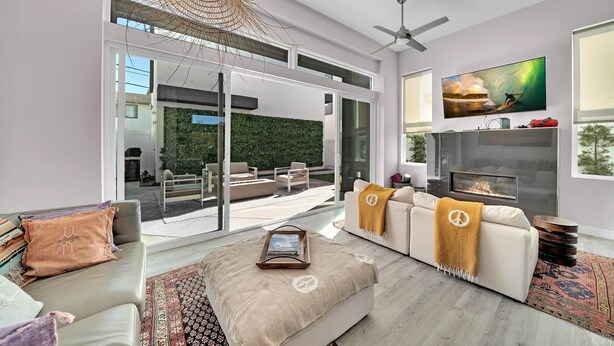 A modern living room with a sofa, coffee table, and a fireplace. There is a television mounted on the wall and a large window providing a view of the outdoors.