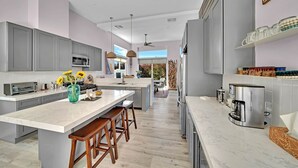 A modern kitchen with grey cabinets, a large island with stools, a coffee machine, and a view to the outdoors.