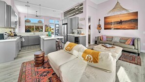 A modern kitchen with grey cabinets, a central island, and a dining area. There is a sofa with cushions and a hanging light fixture.