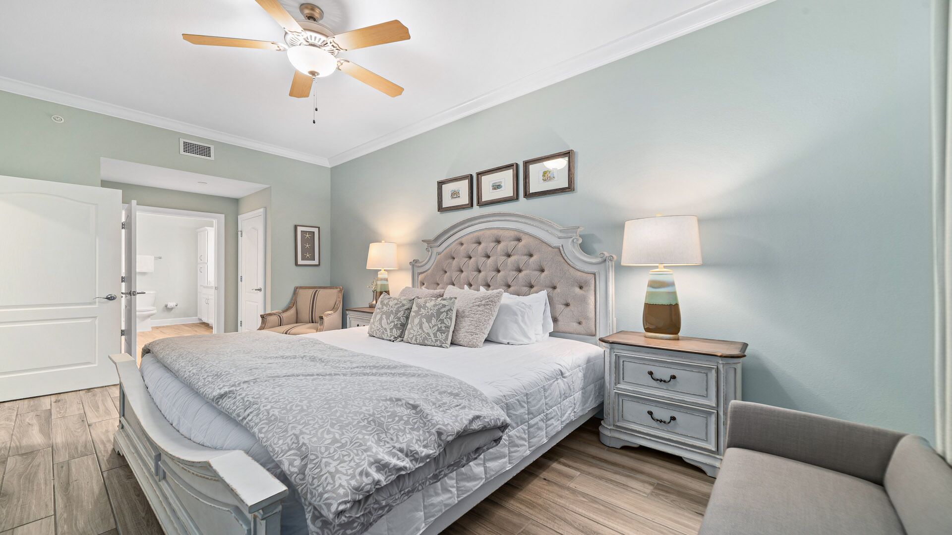 A bedroom with a tufted headboard bed, a bedside table with a lamp, a chair, and a ceiling fan.