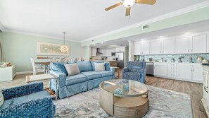 Uma sala de estar ampla com um sofá azul, uma mesa de centro de vidro e um espaço de jantar com mesa e cadeiras brancas.