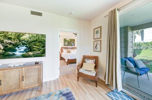 A bedroom with a bed, a TV, a wicker chair, and a balcony with a view of greenery. - Summer Deal Poipu Villa Near Beach with Resort Pool and Hot Tub (Koloa)