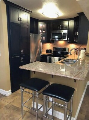 A modern kitchen with dark cabinetry, stainless steel appliances, a marble countertop, and two bar stools.
