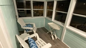 A covered patio area with two white lounge chairs and a small table, featuring a view of the outdoors through large windows. - Cozy Cottage in Snug Harbor, minutes to the beach (pet friendly) (South Kingstown)