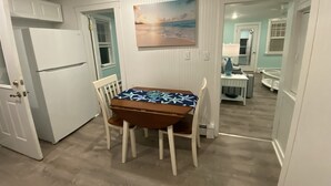 A compact kitchen with a dining table, chairs, a refrigerator, and a painting of a beach scene. - Cozy Cottage in Snug Harbor, minutes to the beach (pet friendly) (South Kingstown)