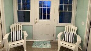 A room with a white door, two windows, and two white chairs with striped cushions. - Cozy Cottage in Snug Harbor, minutes to the beach (pet friendly) (South Kingstown)