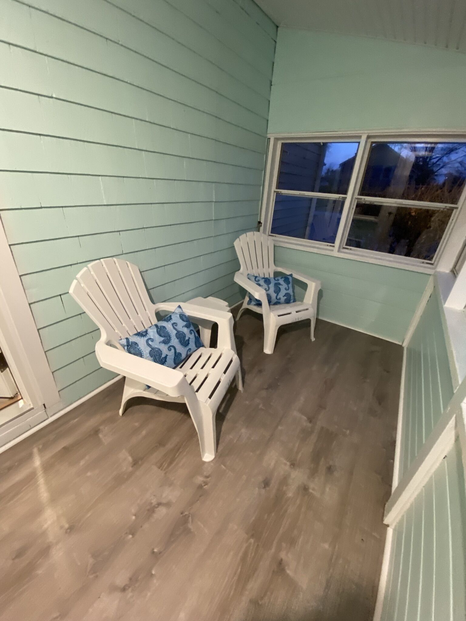 Un porche cubierto con dos sillas Adirondack, revestimiento azul claro y una ventana.
